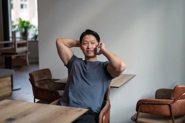 Kendine güvenen genç bir adam kafeteryada telefonla konuşuyor, elleri başının arkasında oturuyor..