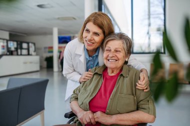Tekerlekli sandalyedeki yaşlı hasta ve klinikte gülümseyen kadın doktor. Kıdemli sağlık hizmetleri.