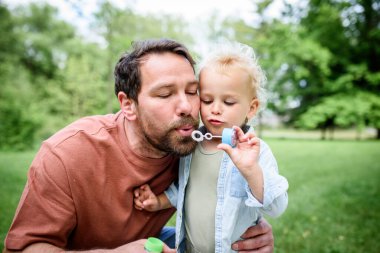 Baba oğul parkta sabun köpüğü üflüyorlar. Bebek şaşkınlıkla kabarcıkları izliyor..