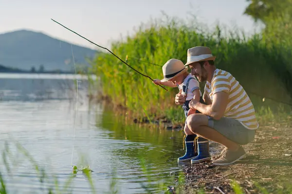 Babasının ve küçük oğlunun, elinde olta tutarken, gölün kenarında dikilirken görüntüsü.