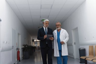 Yönetici doktorla koridorda yürüyor, tabletteki belgeyi gösteriyor. Hastane ayarları.