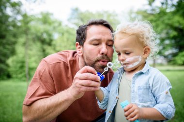 Baba oğul parkta sabun köpüğü üflüyorlar. Bebek şaşkınlıkla kabarcıkları izliyor..