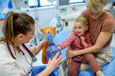 Kadın çocuk doktoru, genç hastaya oyuncak ayının üzerinde oksijen maskesi göstermiş.
