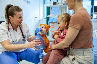 Kadın çocuk doktoru, genç hastaya oyuncak ayının üzerinde oksijen maskesi göstermiş.