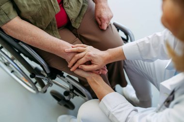 Doktor rahatlatıcı konuşma sırasında tekerlekli sandalyedeki yaşlı kadınla el ele tutuşuyor..