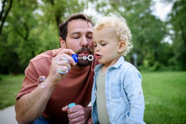 Baba oğul parkta sabun köpüğü üflüyorlar. Bebek şaşkınlıkla kabarcıkları izliyor..