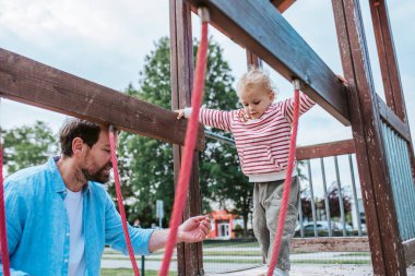 Çocuk, babasının desteğiyle çocuk parkı engelini aşıyor. Hafta sonu aktivitesi..