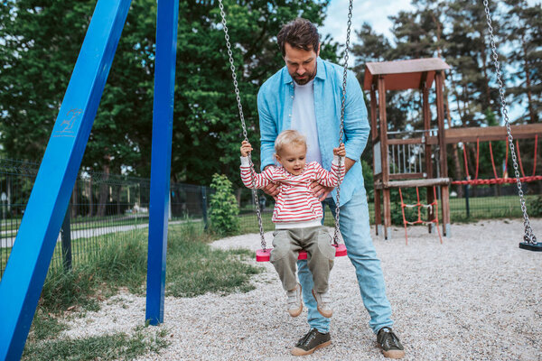 Отец размахивает маленьким сыном на качелях на детской площадке, веселье на выходных.