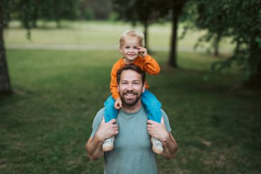Babam parkta yürürken gülümseyen oğlunu omuzlarında taşır.