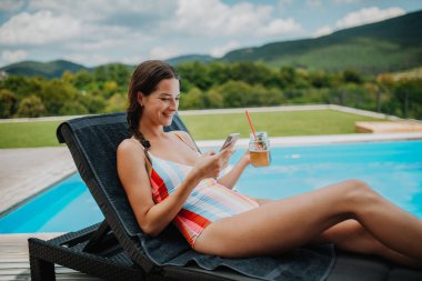 Genç bir kadın havuz kenarında oturuyor, akıllı telefondan kayıyor ve gülümsüyor..