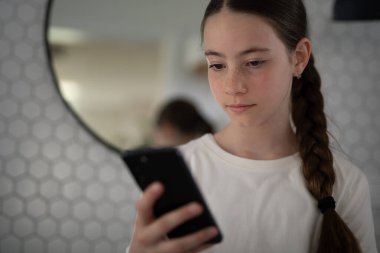 Genç kız banyoda duruyor ve akıllı telefonunu kontrol ediyor, sosyal medyada geziniyor..