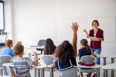 Küçük okul çocukları derste sınıfta masada otururken, el kaldırarak.