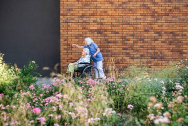 Tekerlekli sandalyedeki yaşlı kadını iten hemşirenin yan görünüşü.