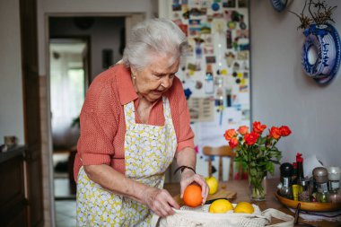 Önlüklü yaşlı bayan evde taze meyve hazırlıyor, elinde de kullanılabilir çanta var..