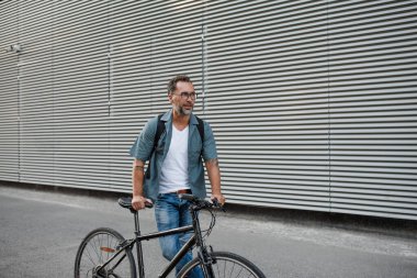 Sokakta bisiklet süren, sırt çantası takan bir banliyö. Kameraya bakıp gülümsüyor..