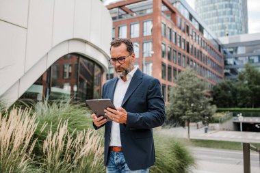 Modern ofis binasının önünde elinde tablet olan ve bir şeyler okuyan bir iş adamı..