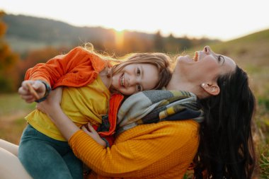 Gülen bir anne ve küçük bir kızı sonbahar doğasında bir yerde oturuyor..