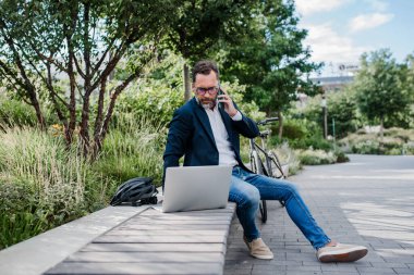 Şehir parkında çalışan erkek yönetici, telefonla konuşuyor ve dizüstü bilgisayarda daktilo yazıyor..