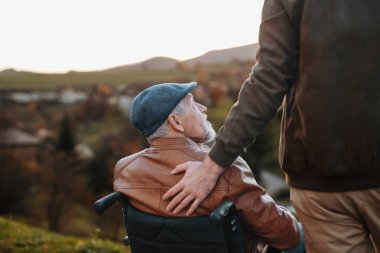 Torun, doğada yürürken büyükbabasını tekerlekli sandalyeye itiyor. Sonbahar mevsimi.