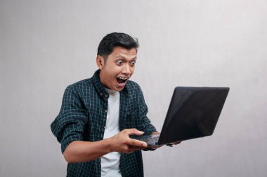 Gingham yeşili tişörtlü bir adam dizüstü bilgisayarını kaldırmış ve şaşırmış bir ifadeyle elinde tutuyor..