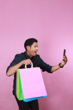 Kareli tişörtlü Endonezyalı adam cep telefonuyla konuşuyor ve pembe arka planda izole edilmiş alışveriş çantaları taşıyor.