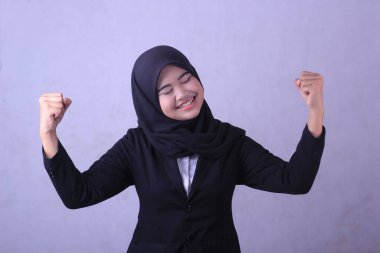 Siyah elbiseli, siyah eşarplı ve kırmızı gülüşlü bir kadın gri bir duvarın önünde duruyor..