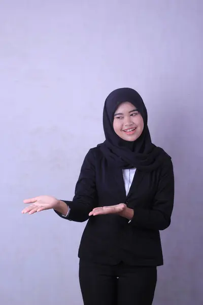A woman in a black suit with a white shirt and black hijab stands in front of a white wall.
