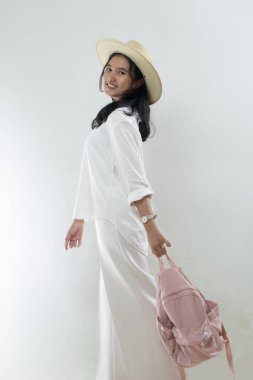 Beyaz gömlekli güzel bir kadının portresi hasır şapka takıyor ve beyaz bir arka planda mutlu bir şekilde yürüyen bir ping çantası taşıyor.