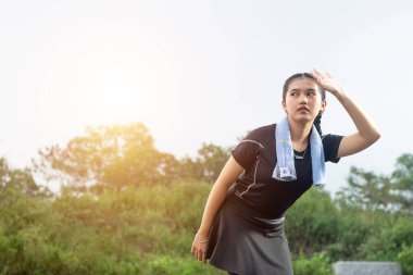 Siyah koşucu kıyafetleri içinde öne eğilmiş genç bir Asyalı kadın terliyor ve eli alnında, öğleden sonra doğa arka planında.