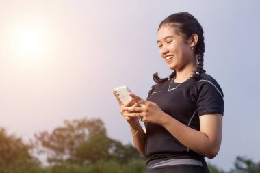 Yan tarafta duran genç bir Asyalı kadın akıllı telefonuyla oynuyor ve siyah koşucu kıyafetleriyle geniş bir gülümseme içinde koşucu kadın, öğleden sonra doğa geçmişi, izole edilmiş negatif bir alan.