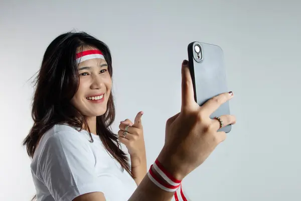 stock image Young Asian Indonesian woman energetically with a headband tied and celebrating Indonesia's independence day while taking a selfie with her cell phone isolated on white background