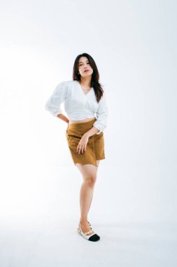 Uzun boylu esmer bir kadının tam boy portresi yalnız başına kameraya bakıyor, beyaz bir ceket ve kahverengi bir şort giyiyor.