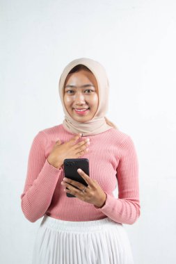 Kameraya gülümseyen ve beyaz renkli siyah cep telefonu uygulamasını kullanan genç kadın, başörtüsü, pembe örülmüş dış elbise ve beyaz bir etek giyiyor..