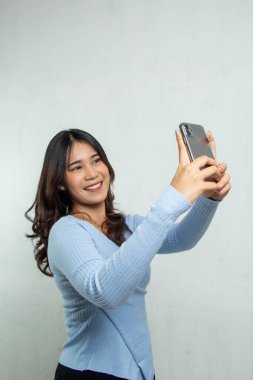 Açık mavi takım elbiseli güzel Asyalı kız cep telefonuyla selfie çekiyor ve mutlu bir şekilde gülümsüyor. Reklamlarda ayakta duran yetişkin kadın.