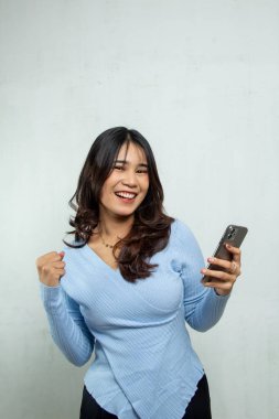 Güzel bir Asyalı kızın yarı vücut portresi başarılı bir açık mavi tarzla heyecanını ifade ediyor ya da cep telefonundan gelen haberlerle ön kameraya bakıyor.