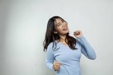 Genç, güzel Asyalı kadın izole edilmiş stüdyonun beyaz duvarında açık mavi bir bluzla dans ediyor.