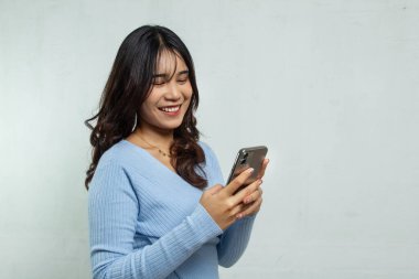 Güzel ve neşeli genç bir kadın elinde akıllı bir telefonla oynuyor. Dar mavi bir bluz ve uzun saçlar giyiyor. İzole edilmiş cep telefonuna bakarken ayakta duruyor..