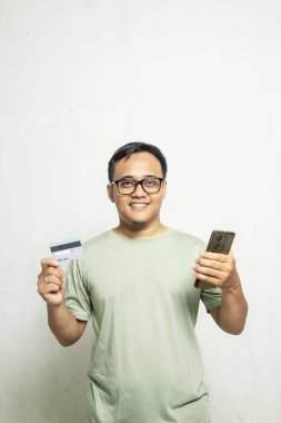 Gözlüklü Asyalı adam kameraya bakıp gülümserken cep telefonu ve kredi kartını beyaz arka planda izole etmiş.