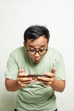 Gözlüklü Asyalı bir Endonezyalı 'nın portresi akıllı telefonuyla oyun oynarken ya da eğlenceli bir şey hakkında haber yaparken şaşırıyor. Yarı vücut adamı ve adaçayı yeşili gömlek.