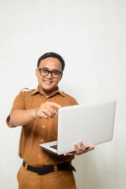 Asyalı Endonezya 'lı öğretmen elinde beyaz dizüstü bilgisayarla kameraya mutlu bir gülümsemeyle bakıyor. Beyaz arka planda eski haki rengi takım elbiseli, Endonezyalı devlet memuru
