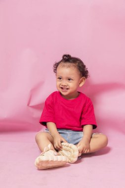 portrait of an 18 month old curly haired girl sitting laughing out loud while looking away isolated on pink clipart