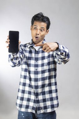 Kareli tişörtlü bir adam beyaz stüdyo duvarında siyah ekranlı telefonu parmağıyla gösteriyor.