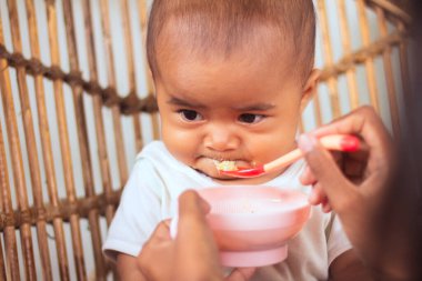 Asyalı bir bebek, annesinin eliyle beslenirken sandalyede surat asmayı reddediyor.