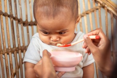 Asyalı bir bebek, annesinin eliyle beslenirken sandalyede somurtmayı reddediyor, kızgın yüzünü kapatıyor.