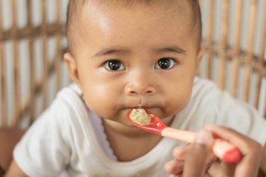 Kimliği belirlenemeyen bir kadının bebeğini evde emzirdiği fotoğraf. Bebek bir rattan üzerine oturur ve ona nazikçe bakar..