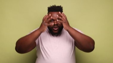 Şaşkın Afro-Amerikalı bir adam pembe tişört giyiyor ve kameraya büyük gözleriyle bakıyor, ağzı açık ve yüzüne dokunuyor. Açık yeşil arka planda kapalı stüdyo çekimleri.