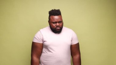 Sinirli, stresli, Afro-Amerikan bir adam pembe tişört giyiyor. Yumruklarını öfkeyle sıkmış, nefret dolu, sinirli ve agresif. Açık yeşil arka planda kapalı stüdyo çekimleri.