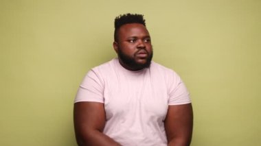 Pembe tişört giyen Afro-Amerikalı bir adam yoga pozisyonunda durup rahatlamaya ve sakinleşmeye çalışıyor, avuç içlerini bir arada tutuyor. Açık yeşil arka planda kapalı stüdyo çekimleri.