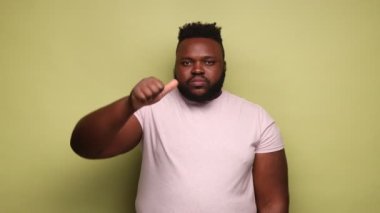 Hoşnutsuz Afro-Amerikalı bir adam pembe tişört giyip baş parmağını aşağı gösteriyor, olumsuz duygularını ifade ediyor, nefret ediyor, suratını asıyor. Açık yeşil arka planda kapalı stüdyo çekimleri.