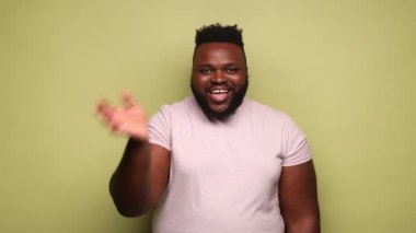 Arkadaş canlısı, Afro-Amerikan bir adam seni selamlıyor, el sallıyor, selam veriyor, seni gördüğüne seviniyor, dişlek gülümsemesiyle kameraya bakıyor. Açık yeşil arka planda kapalı stüdyo çekimleri.